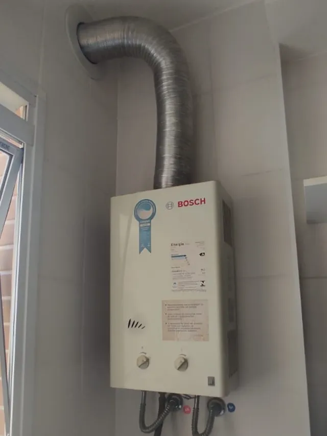 Aquecedor a gás Bosch mecânico instalado em área de serviço, com chaminé até a parede e ligações flexíveis de água e gás
