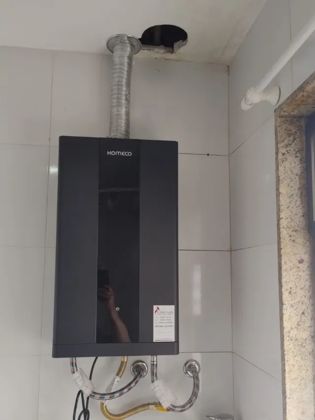 Aquecedor a gás Komeco digital preto instalado em área de serviço, com chaminé no teto e etiqueta de revisão da Configás