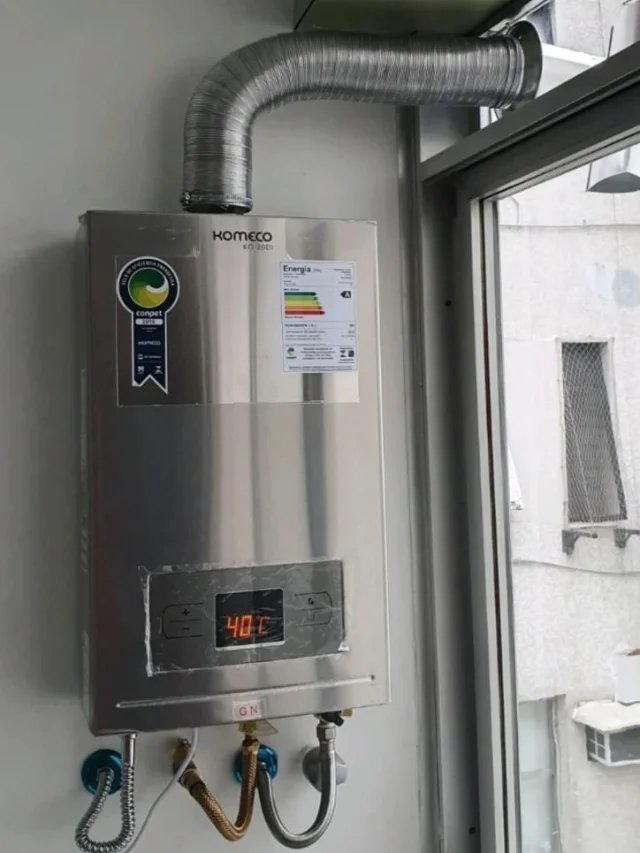 Aquecedor a gás Komeco KO 20DI inox digital marcando 40 °C, com chaminé até a janela e ligações de gás natural